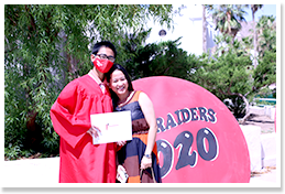 Graduate wearing a graduation robe posing with an adult in front of a Red Raiders 2020 sign